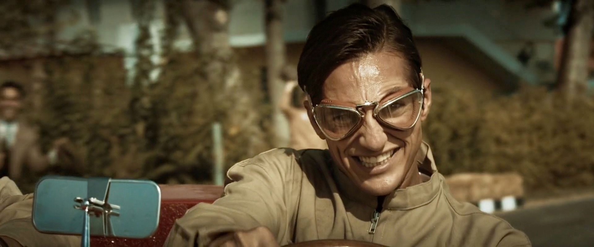 A film frame: a driver in racing goggles grins over the wheel of an open-top car, a chrome wing mirror in the foreground.