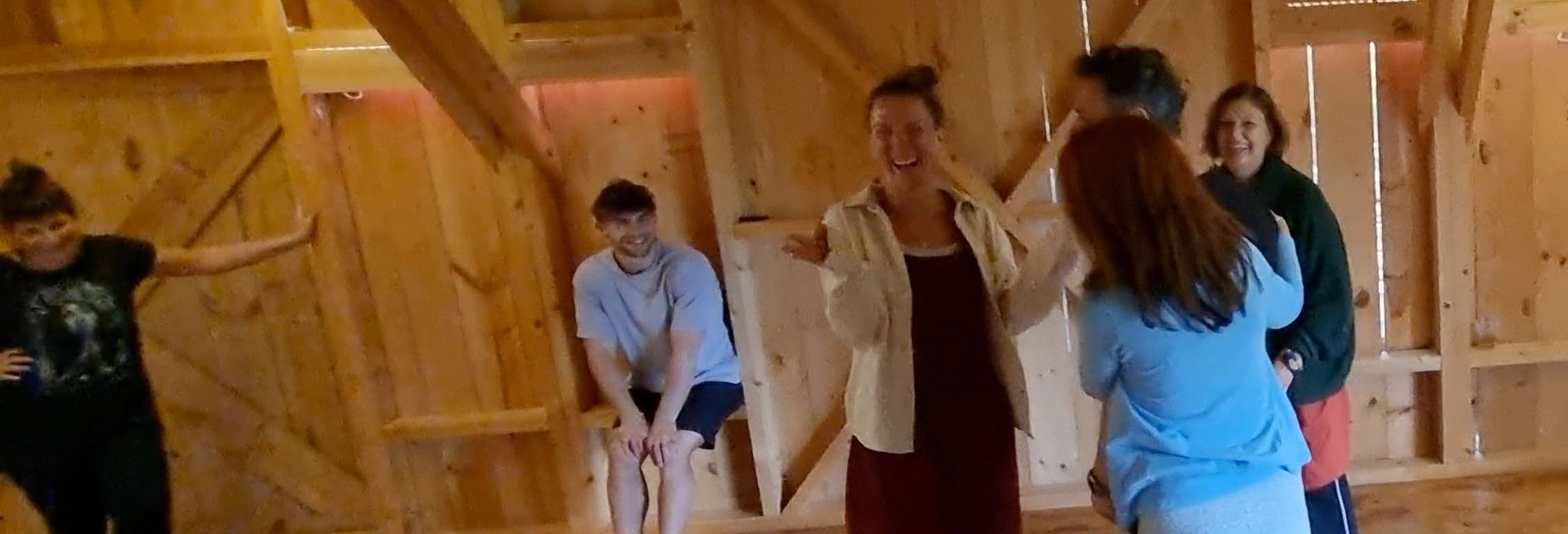 A group of course participants laughing together in a pale timber hall: a woman in the middle brings a hand to her face while the others watch her.