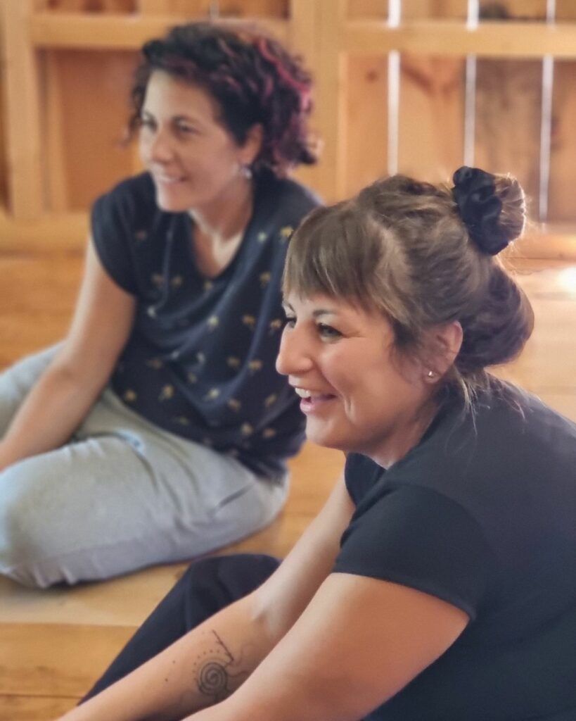 Two course participants sitting on a wooden floor, smiling as they watch an exercise off to one side.
