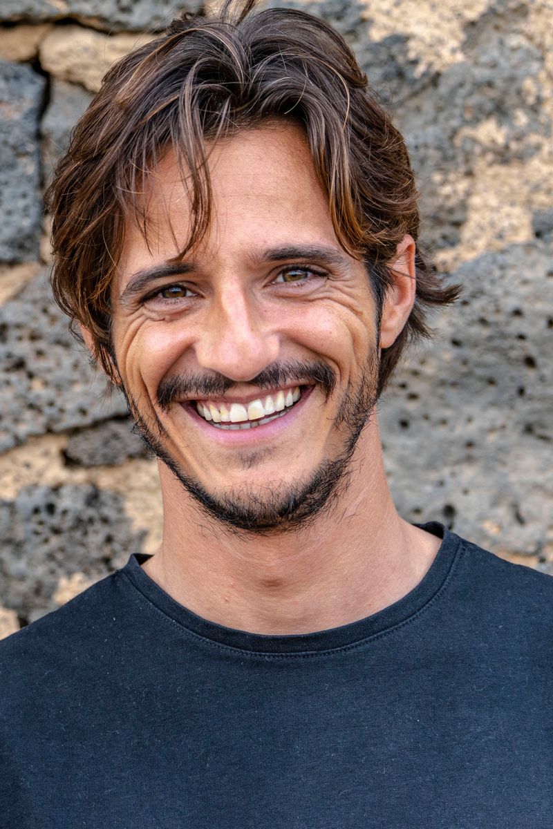 Matteo Leoni smiling at the camera, head and shoulders, wavy brown hair and a short beard, in a dark T-shirt in front of a wall of dark volcanic stone.