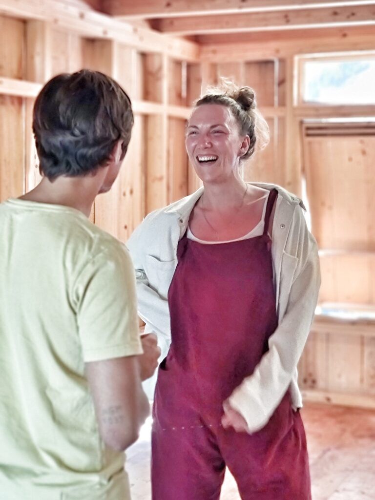 A course participant in dusty-pink dungarees laughing out loud while facing another participant, in a hall lined with pale timber.