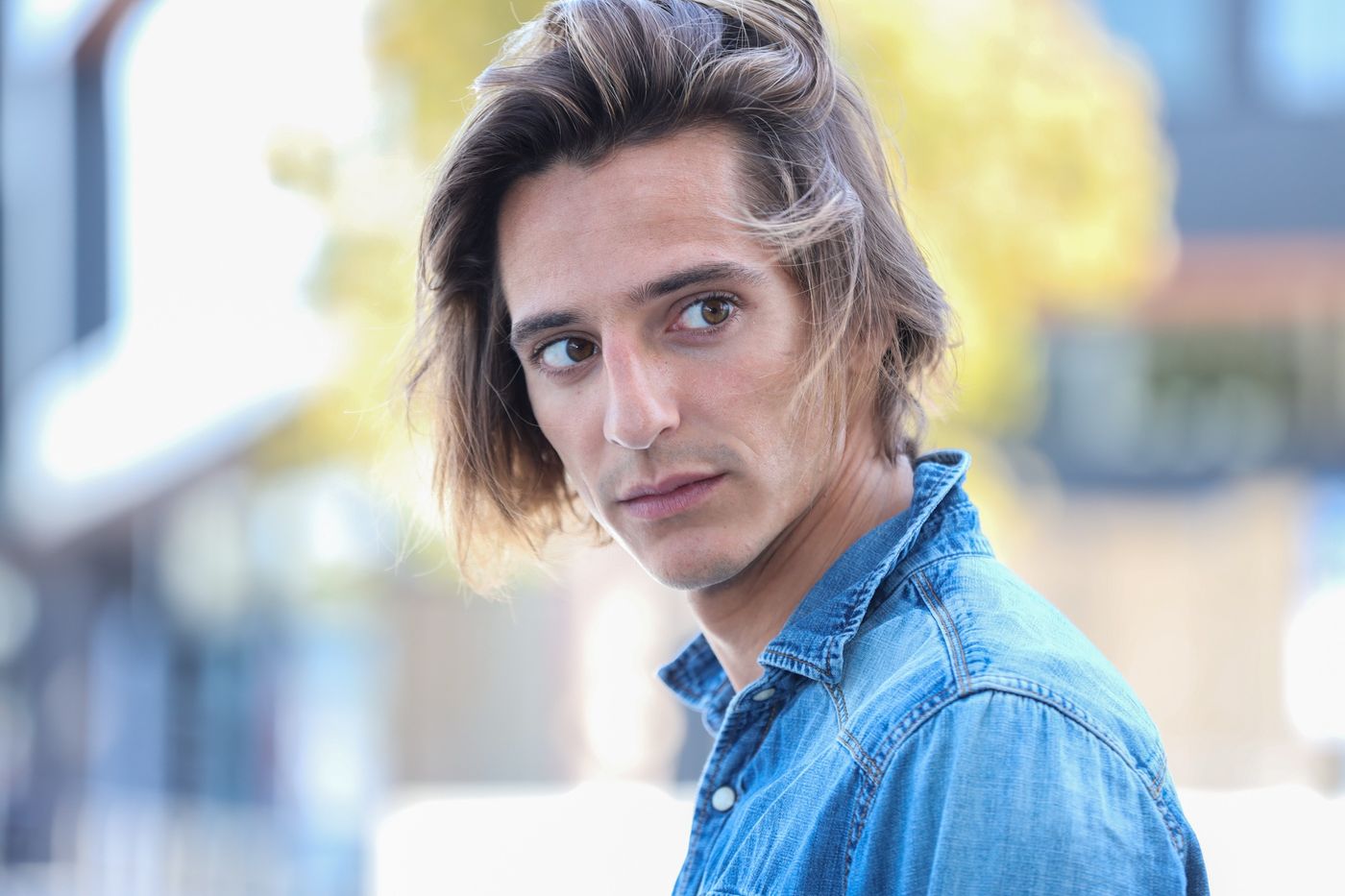 Matteo Leoni in a denim shirt, hair blown across his face, glancing back over his shoulder on a street that is out of focus behind him.
