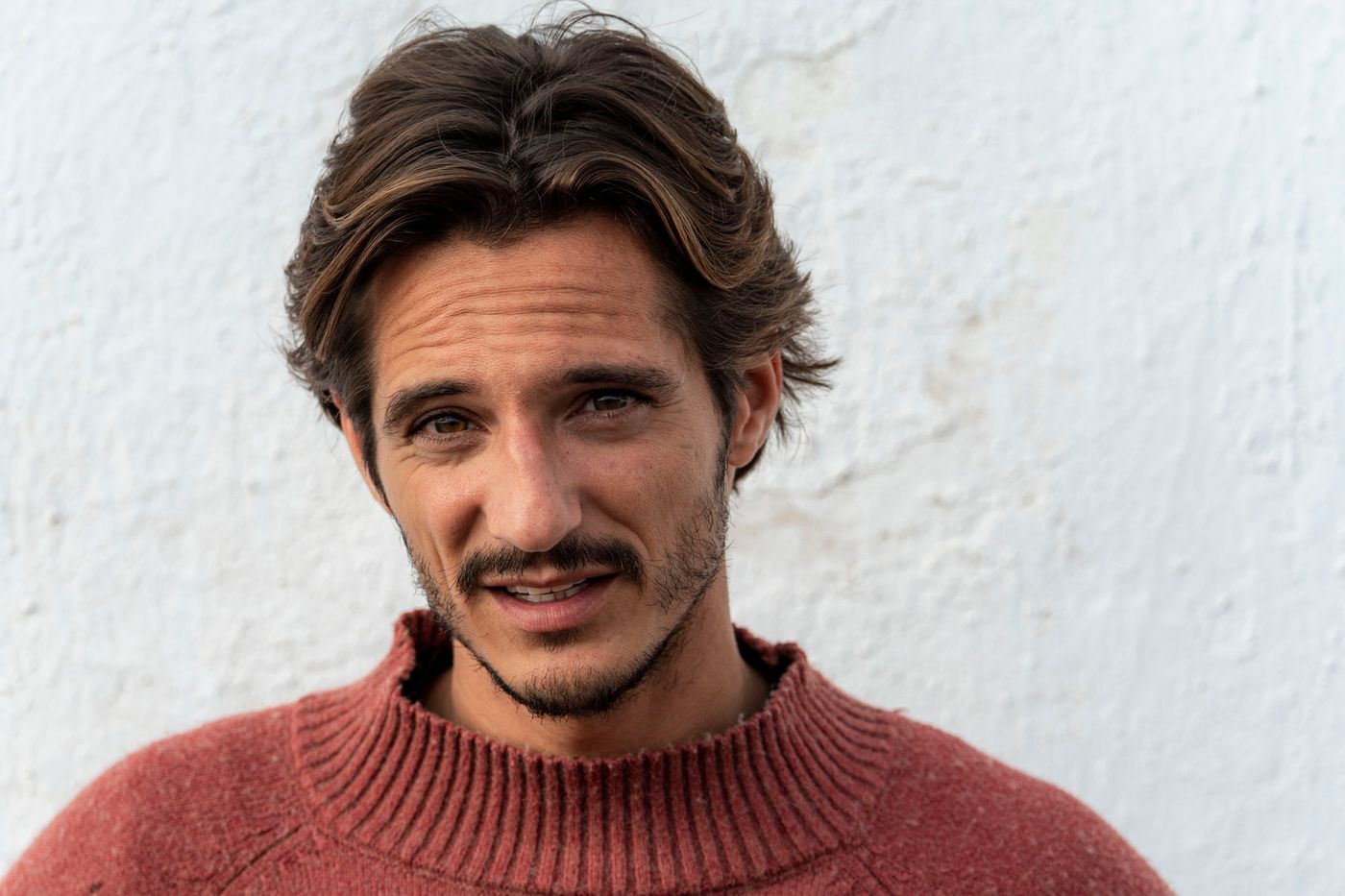 Matteo Leoni smiling with his mouth half open, loose wavy hair, in a rust-red jumper against a white wall.