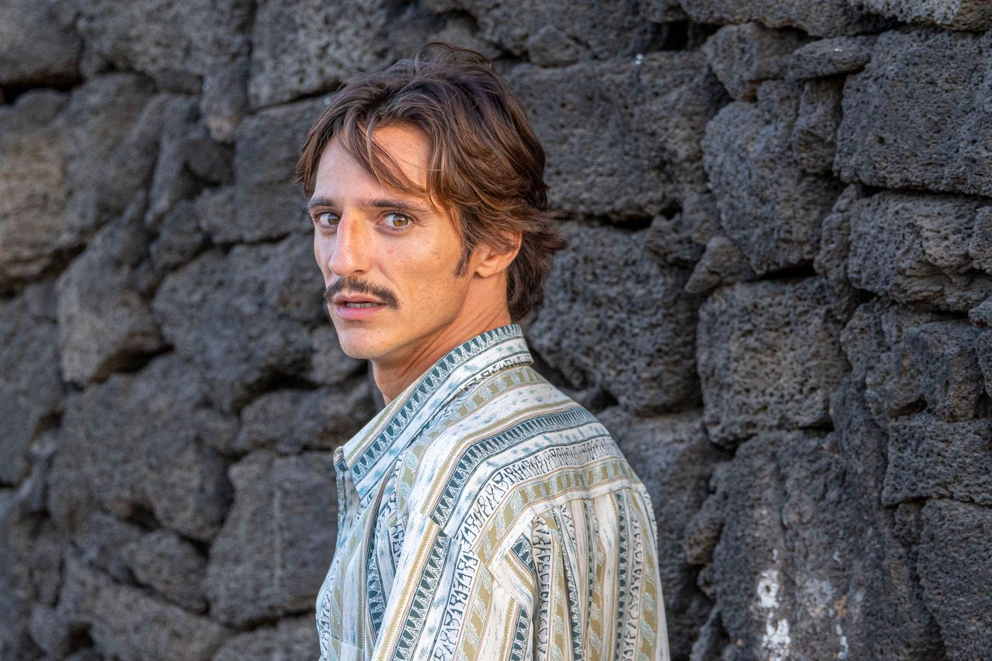 Matteo Leoni in a patterned shirt, turned to one side and looking past the camera, in front of a wall of dark volcanic stone.
