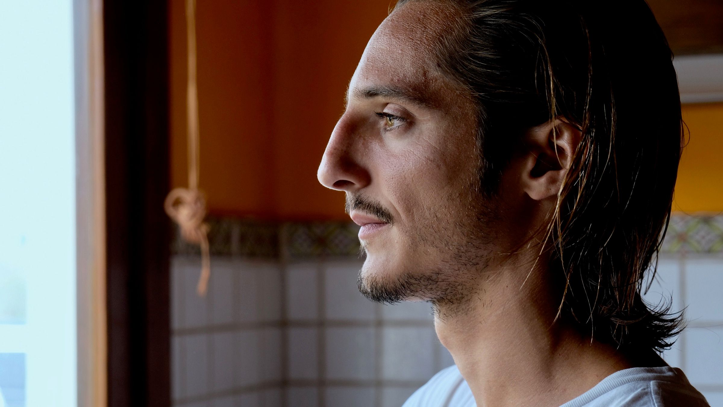 Matteo Leoni in profile, hair pulled back, in a white T-shirt, looking out of a window in a room with tiled walls and warm light.