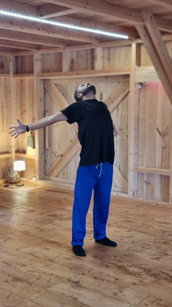 A course participant standing in the middle of a wooden hall in socks, arms stretched out wide and head tipped back.