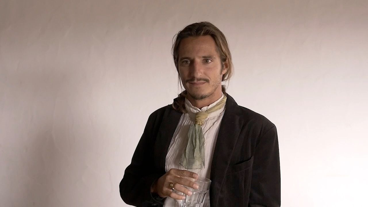 The same character, turned towards the camera, holding a cut-glass tumbler at chest height.