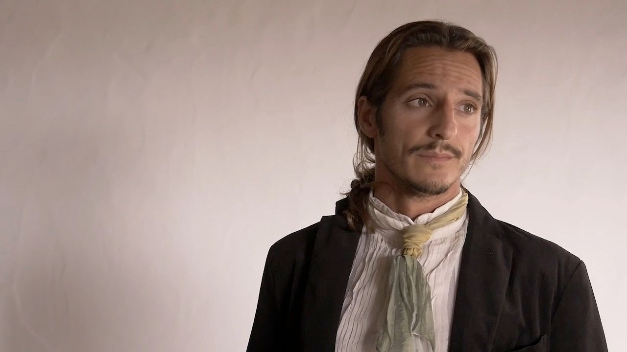 A film still: a man in a dark jacket, a white shirt and a pale green cravat, hair tied back, looking off to one side against a bare wall.
